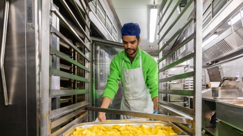 Man in hair net and apron loading tray of potatoes into industrial oven