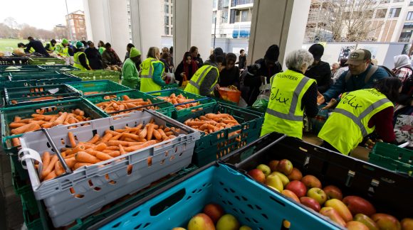 Queue of people receiving food from group of people wearing high-vis and dozens of crates of fresh fruit and veg